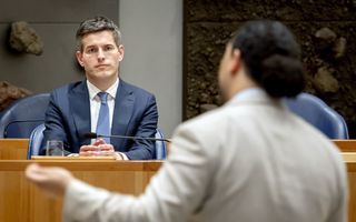 Man in pak zit in kabinetsvak van de Tweede Kamer. Tegenover hem, bij de interruptiemicrofoon, staat een Kamerlid in een licht kostuum.