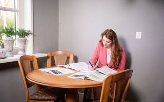 Een meisje met donkerbruin lang haar en een roze blazer zit aan een houten tafel met één poot. Ze maakt notities in een schrift. Om haar heen liggen studieboeken geopend.