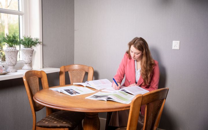 Een meisje met donkerbruin lang haar en een roze blazer zit aan een houten tafel met één poot. Ze maakt notities in een schrift. Om haar heen liggen studieboeken geopend.