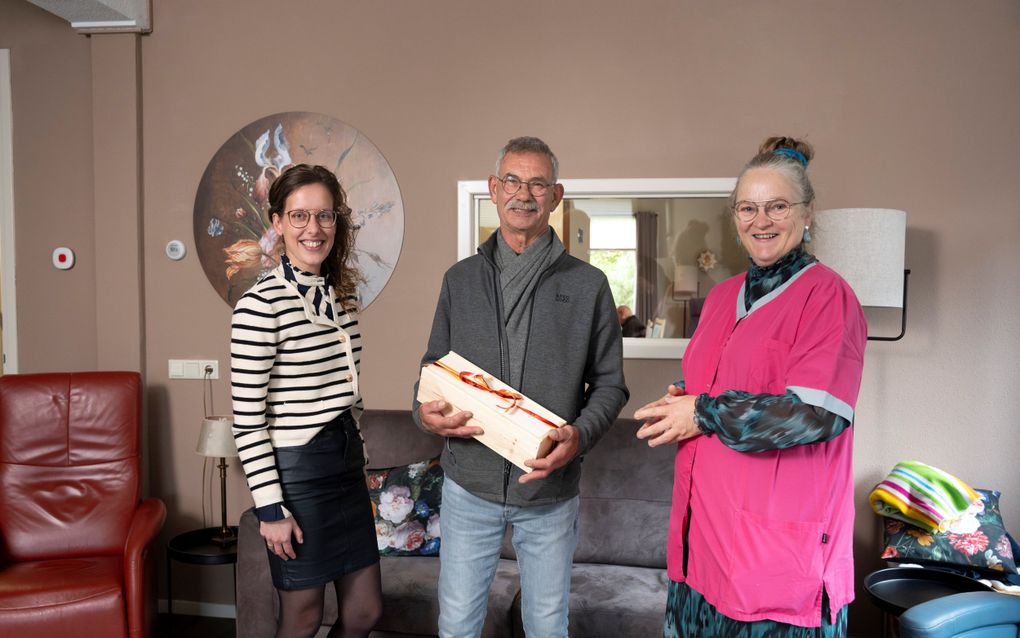 Elisabeth van Vuuren (r.) geeft een notenpakket aan vrijwilliger Mark Mulder (m.). Activiteitencoach Verie de Boer-Mulder (l.) haalde Mulder met een smoes binnen zodat hij verrast kon worden. beeld Niek Stam