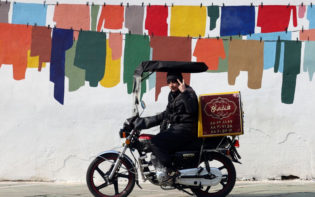 Een Iraanse man laat het overwinningsbord zien terwijl hij naast een muurschildering rijdt in een straat in Teheran, Iran. beeld EPA, Abedin Taherkenareh