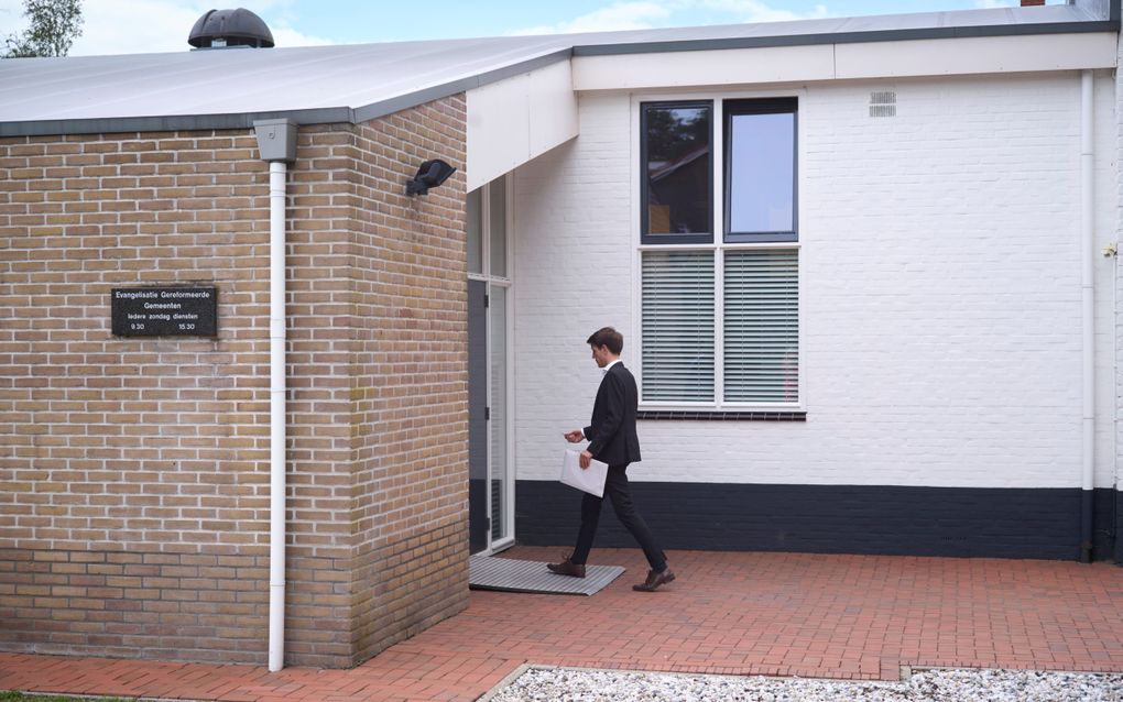 Een jongeman loopt naar de ingang van een gebouw; aan de muur van het gebouw hangt een bord met ”Evangelisatie Gereformeerde Gemeenten. Iedere zondag diensten: 9:30, 15:30