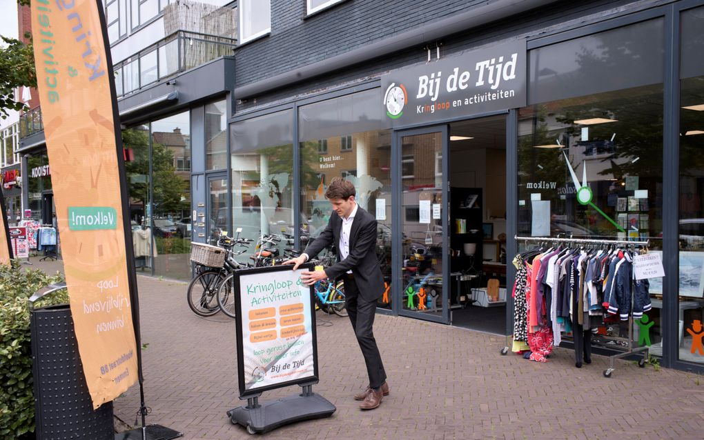 Een jongeman in tweedelig pak zonder stropdas zet een bord op de stoep voor een kringloopwinkel. Op de gevel van de winkel staat: Bij de Tijd; kringloop en activiteiten