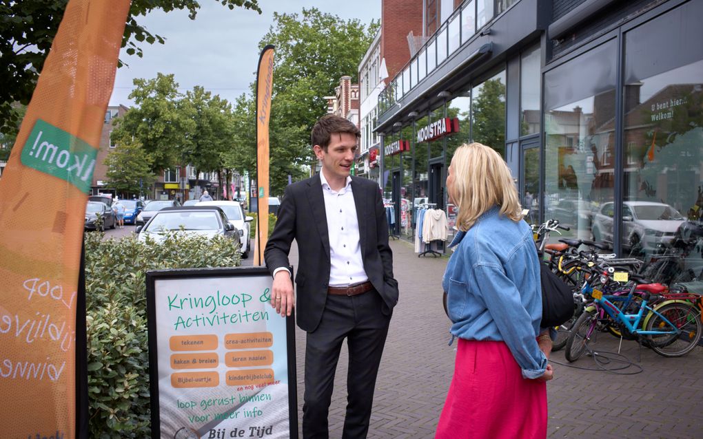 Een jongeman in tweedelig pak zonder stropdas staat op straat te praten met een vrouw met een spijkerjasje en een felroze rok
