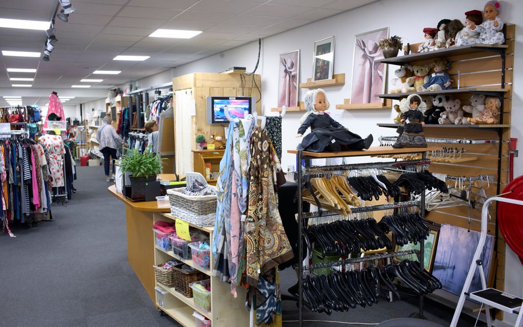 Interieur van een kringloopwinkel, met schappen vol kleding en op de voorgrond wat poppen en kleurrijke doeken; in de achtergrond zijn twee klanten te zien