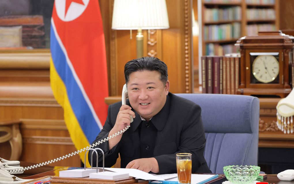 Een man in pak zit lachend achter een bureau aan de telefoon. 