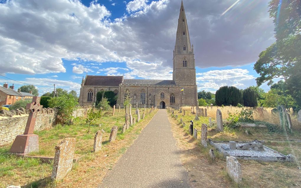 Geasfalteerd pad tussen grafstenen. Aan het einde van het pad staat een stenen kerkgebouw met hoge toren die afsteekt tegen de wolkenlucht.