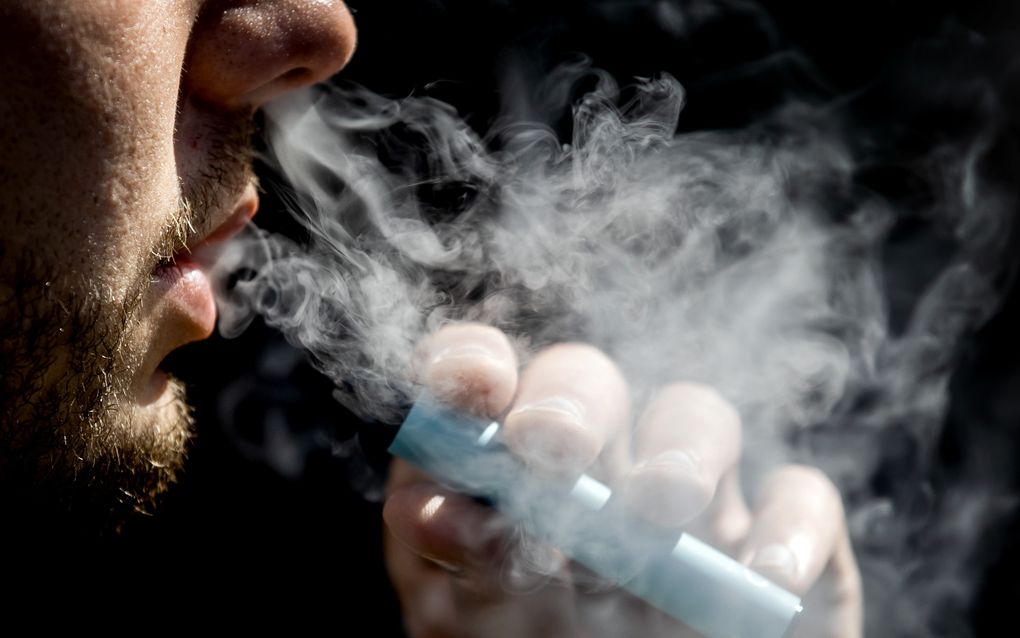 Een bebaarde man houdt een vape vast, een flinke rookwolk komt uit zijn mond.