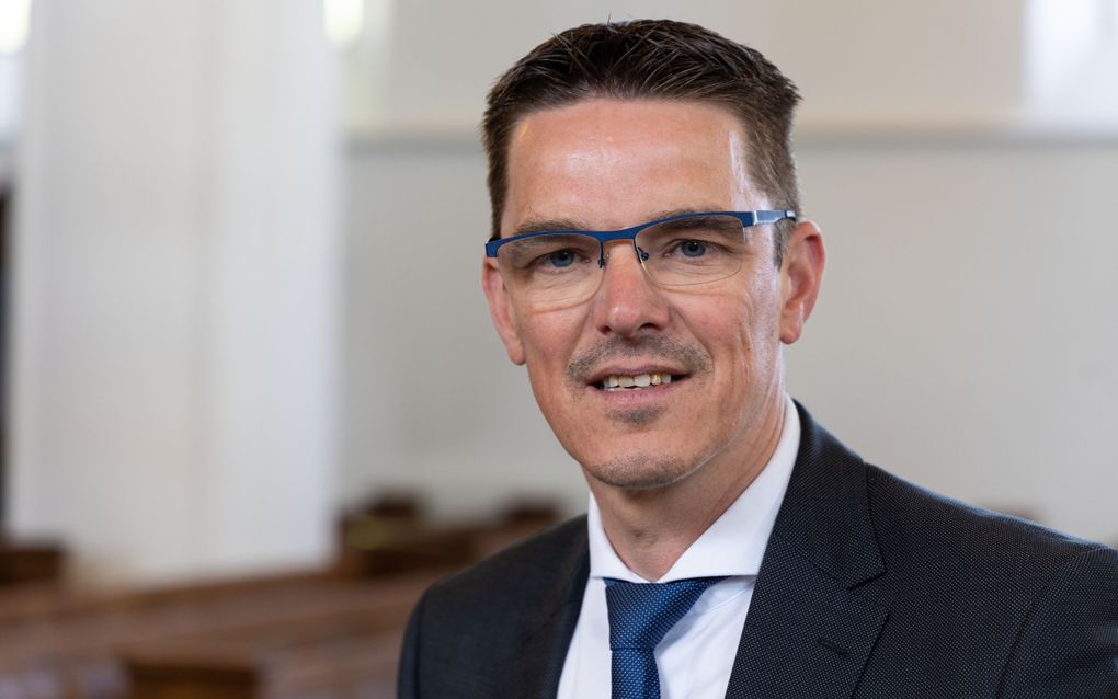 Dominee Van den Noort, een man met zwart haar, bril op en gekleed in donker kostuum met blauwe stropdas.