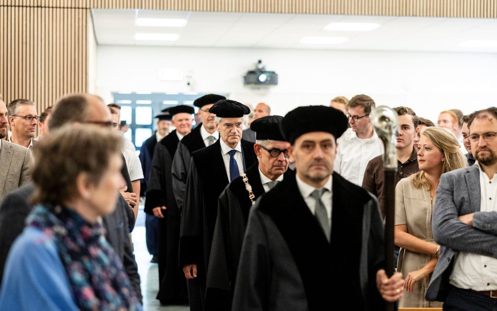 Een rij mannen in zwarte toga’s met zwarte baretten op. Zij lopen door een gangpad een zaal binnen. Links en rechts staan belangstellenden.