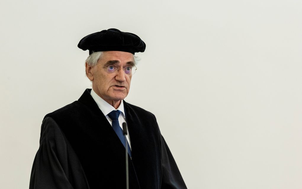 Man in zwarte toga, met zwarte baret, houdt voordracht.