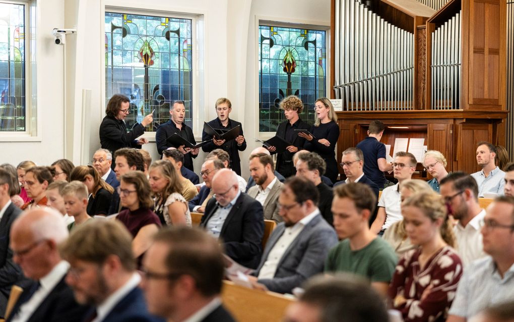 Een zaal met mensen, gezeten in kerkbanken. Achterin een pijporgel en een klein kamerkoor dat staat te zingen.