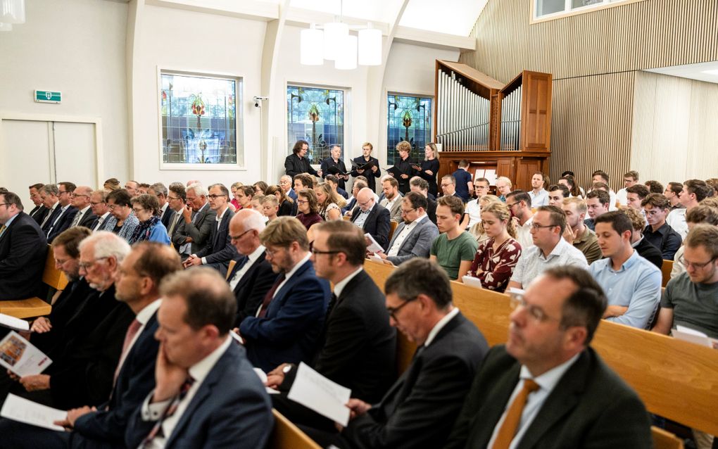 Een zaal met mensen, gezeten in kerkbanken. Achterin een pijporgel en een klein kamerkoor dat staat te zingen. 