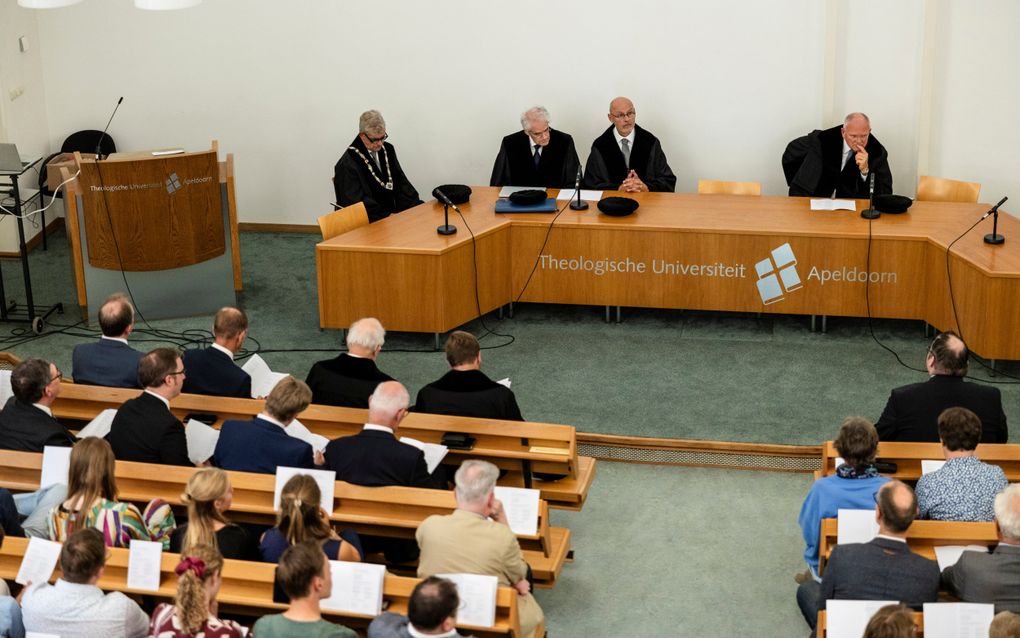 Vier mannen in toga achter een bestuurstafel. Voor hen een zaal vol mensen.