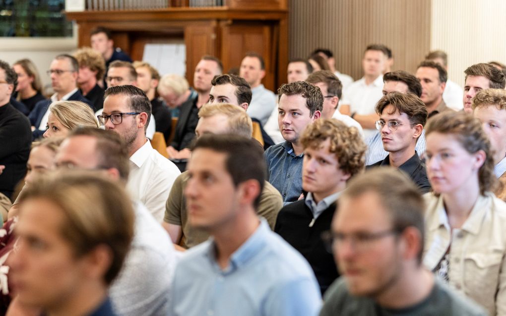 Luisterende gezichten, van jonge mannen en vrouwen, in de zaal van een aula.