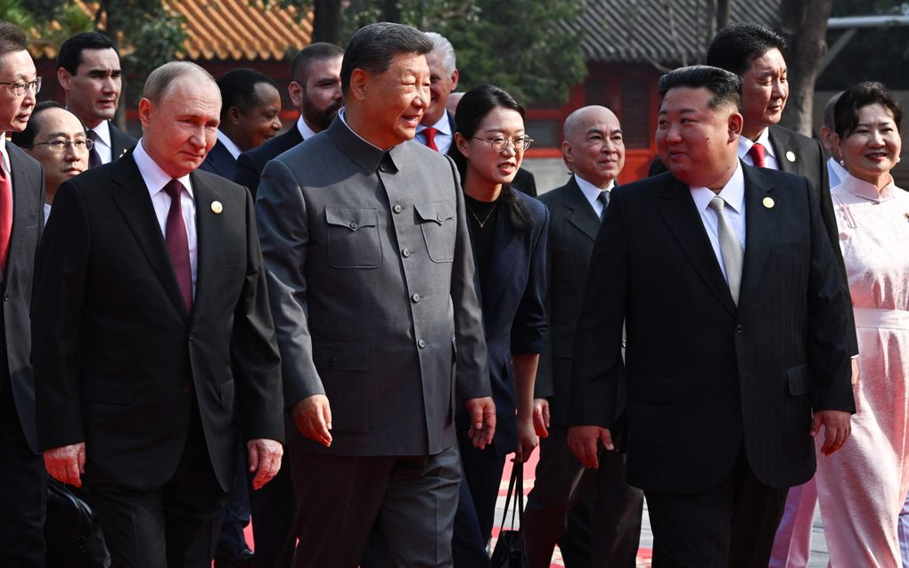 Een man in grijs pak, geflankeerd door twee mannen in donker pak. Achter hen een gevolg. 