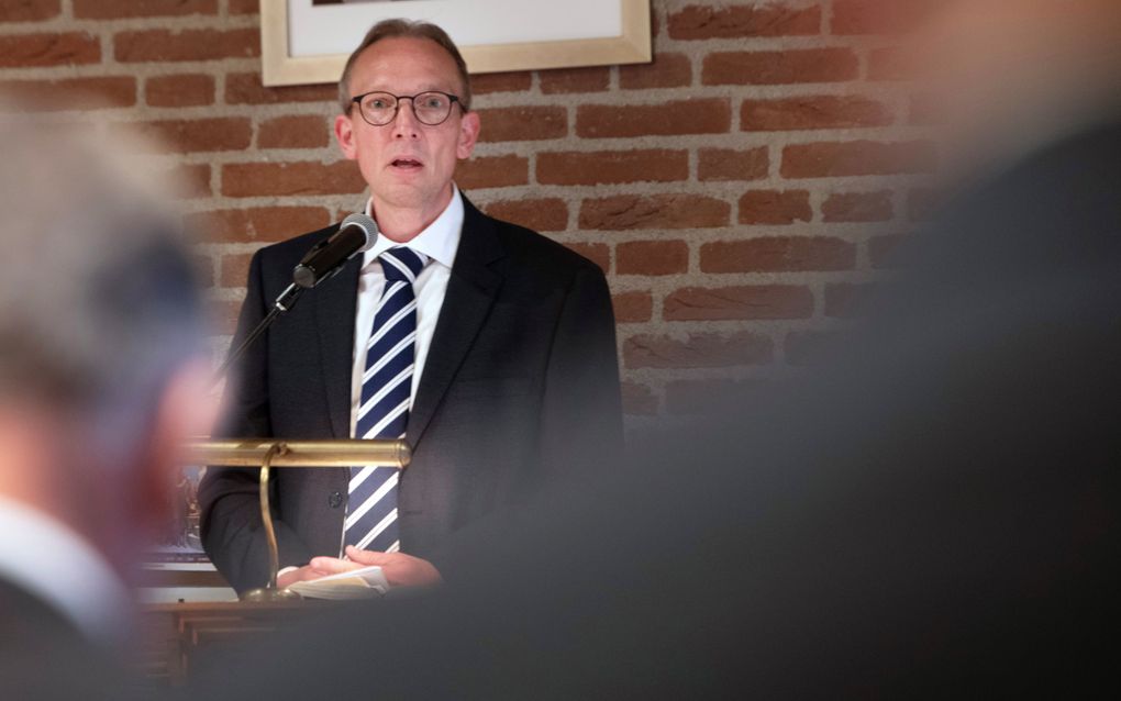 Man met bril, in donker pak, met gestreepte stropdas, achter de katheder.