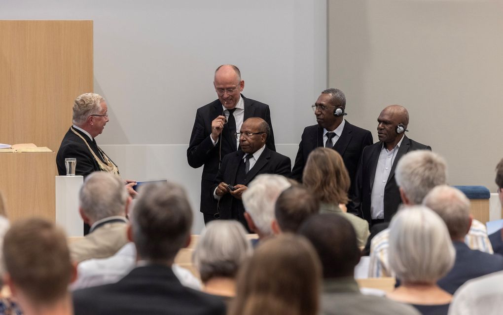 Een man in zwart pak spreekt een andere man in zwart pak toe. Er staan drie zwarte mannen in zwarte pakken bij en op de voorgrond zie je hoofden van mensen van achteren.