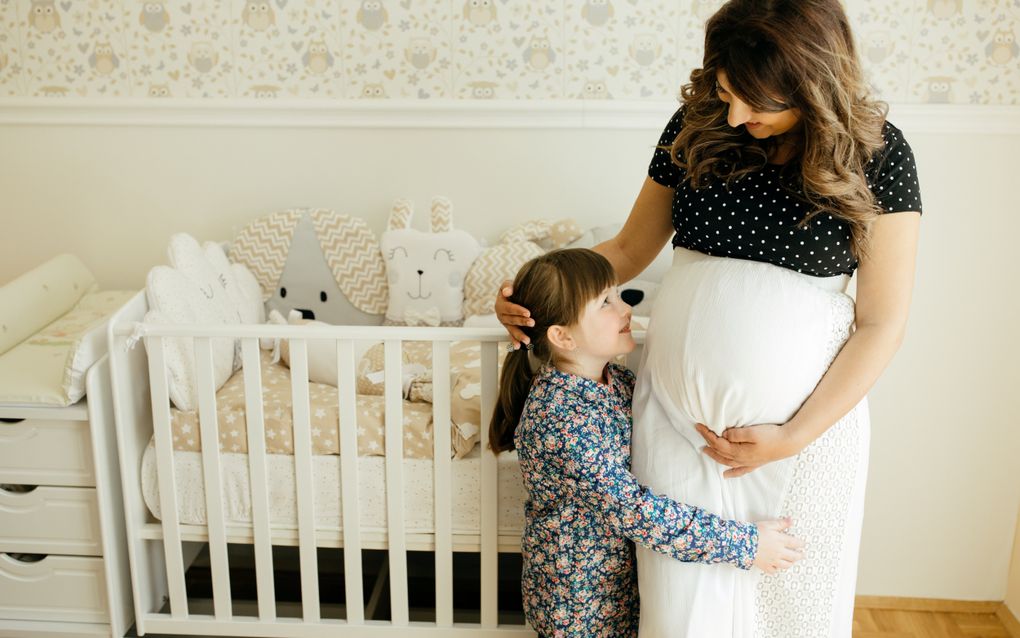 Zwangere vrouw met witte rok en blauwe gestippeld shirt staat in een babykamer bij een ledikantje. Haar dochter, met bloemetjesjurk en paardenstaart, staat bij haar. De vrouw legt haar hand op het hoofd van het meisje.