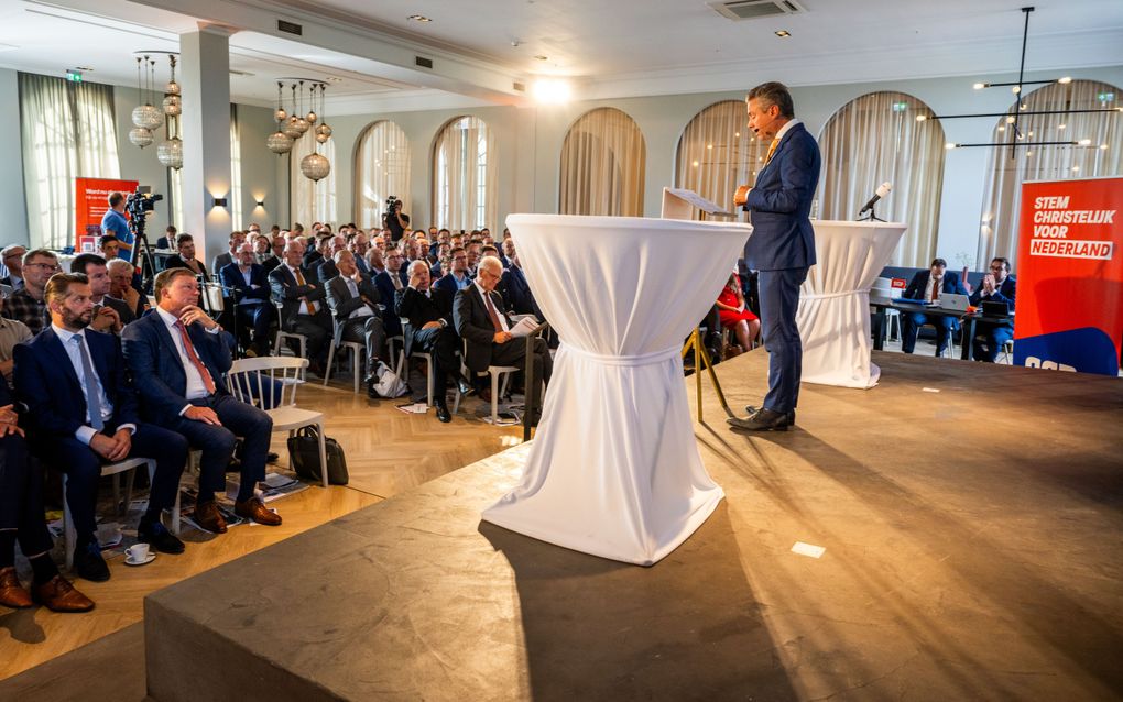 Podium van opzij gezien met een man achter een wit overtrokken statafel, voor hem een groep luisterende mannen.