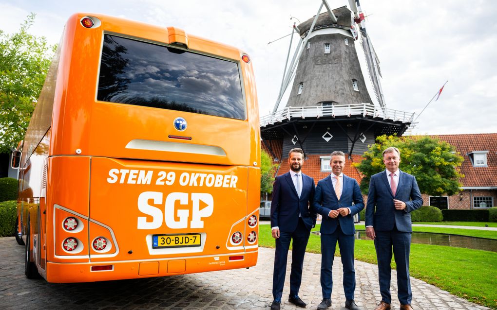 Mannen in pak naast een oranje touringcar en op de achtergrond een molen.
