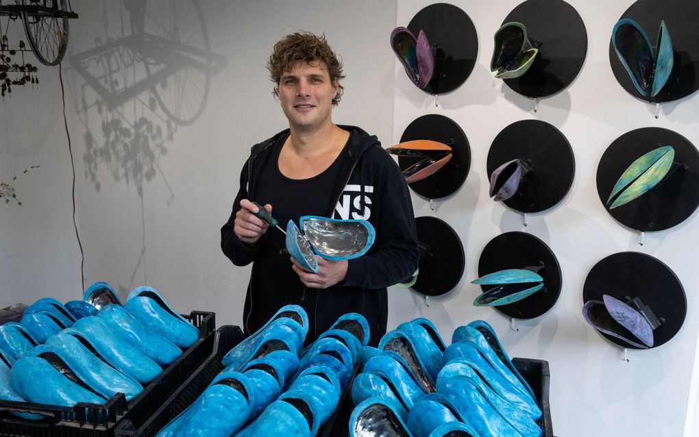 Man met een opengeslagen mossel in zijn handen bij een tafel met nog meer mosselen, en aan de muur achter hem eveneens mosselen.