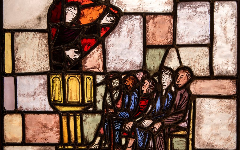 Glas-in-loodraam met Augustinus op een kansel, een hand omhoog. Mensen op stoelen luisteren naar hem.