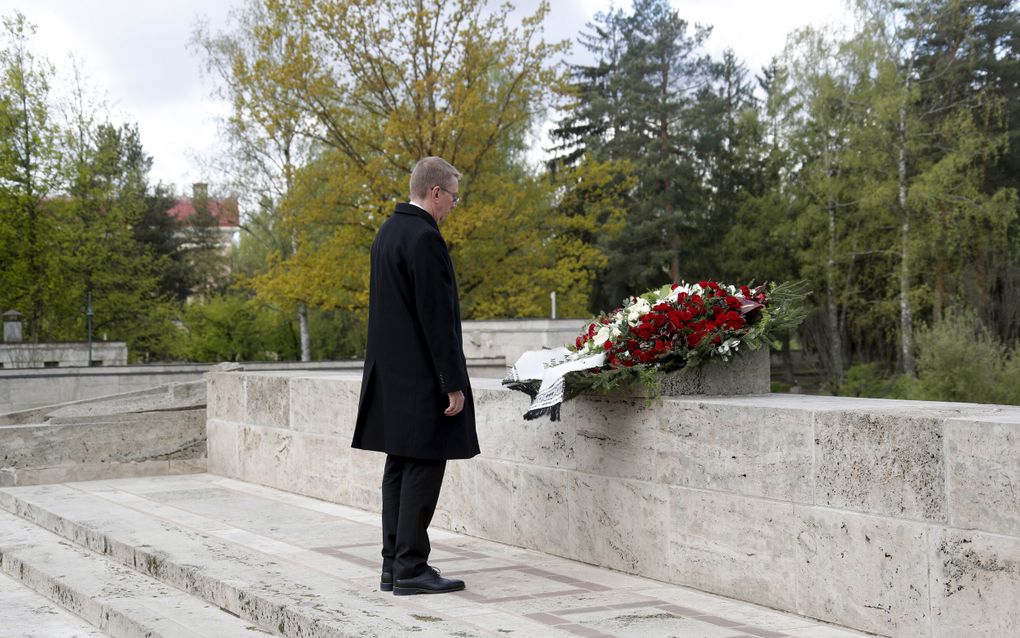 Man in donkere kleding staat voor bloemenkrans die op een stenen muur ligt.