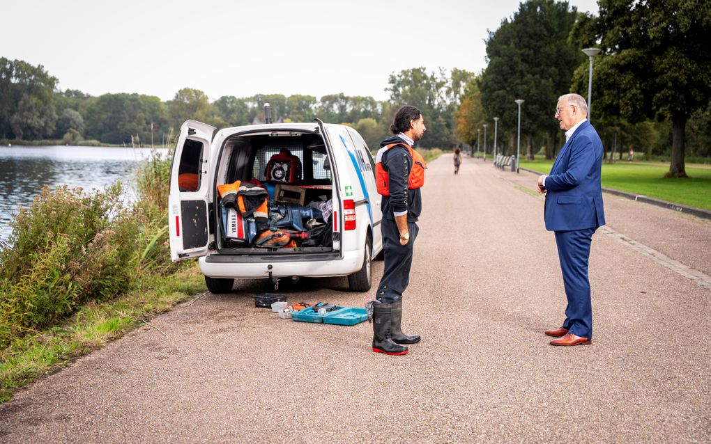 Man praat met een man die een duikvest draagt. Achter hen staat een busje met open deuren, waarin duikattributen liggen.