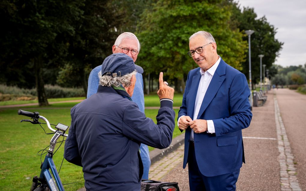 Man praat met twee voorbijgangers. Hij luistert naar een man die een petje draagt en een vinger opgeheven heeft.