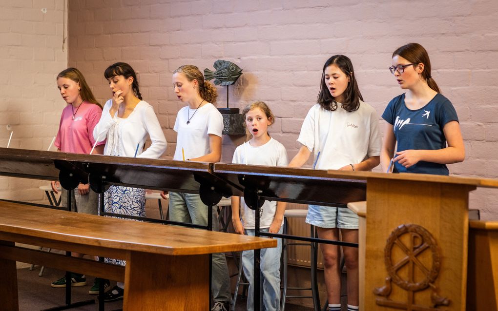 Zes meisjes staan achter een houten lessenaar en zingen. Een van hen kijkt opzij. 