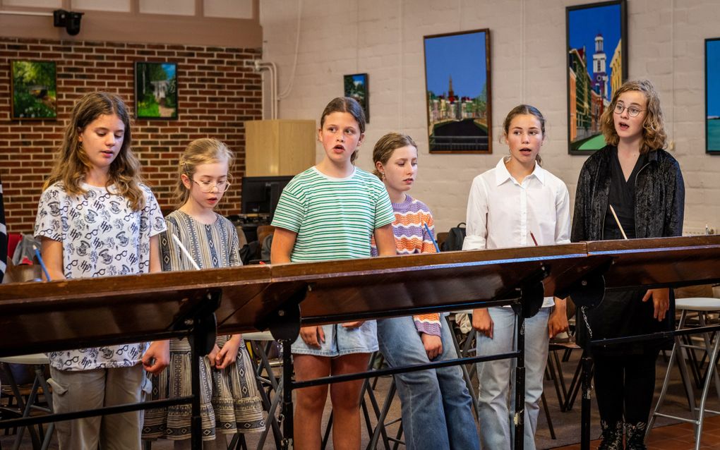 Zes meiden staan achter lessenaars en zijn aan het zingen. Drie meiden kijken opzij naar de dirigent. 