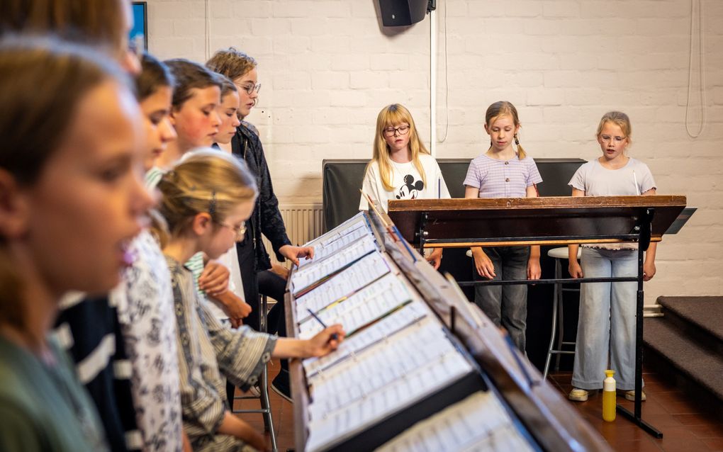 Een groep meiden staat achter lessenaars en zingt. Drie meiden kijken met het gezicht deze kant op. De rij meiden links is vaag in beeld gebracht. 