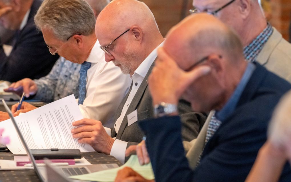 Rij mannen achter een vergadertafel. Ze schrijven of bladeren geconcentreerd in stukken.