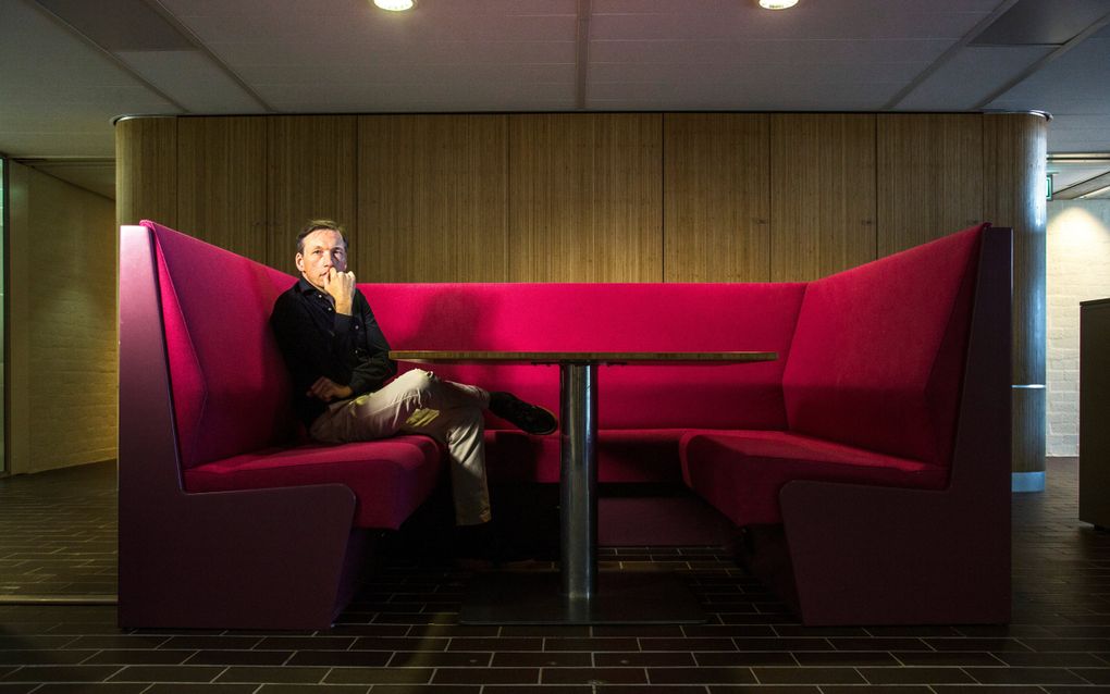 Man van middelbare leeftijd met lichte broek en donkerblauw overhemd zit op een rode bank die in een U-vorm loopt, met een tafeltje in het midden. De man heeft zijn ene hand voor zijn mond, en kijkt peinzend naar de fotograaf