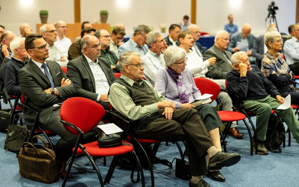 Een zaal met zo’n tachtig mensen, zittend op stoelen, in luisterhouding.