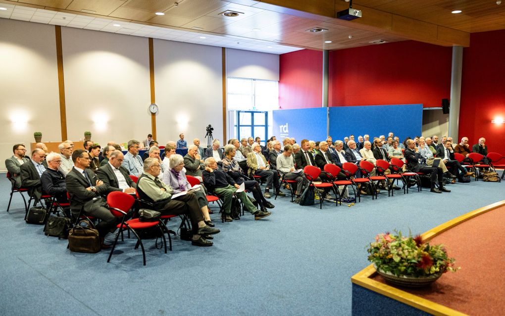 Een zaal met zo’n tachtig mensen, zittend op stoelen, in luisterhouding.