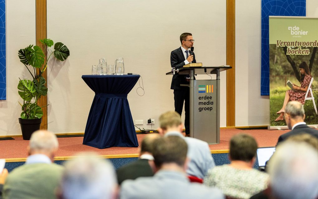 Man in donker pak, op een podium. Voor hem een zaal met luisteraars.
