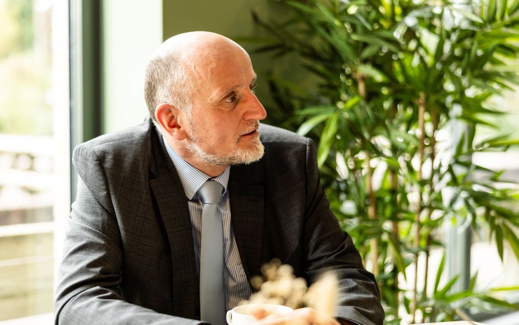 Man in donker pak, met grijs baardje, zittend aan een tafel.