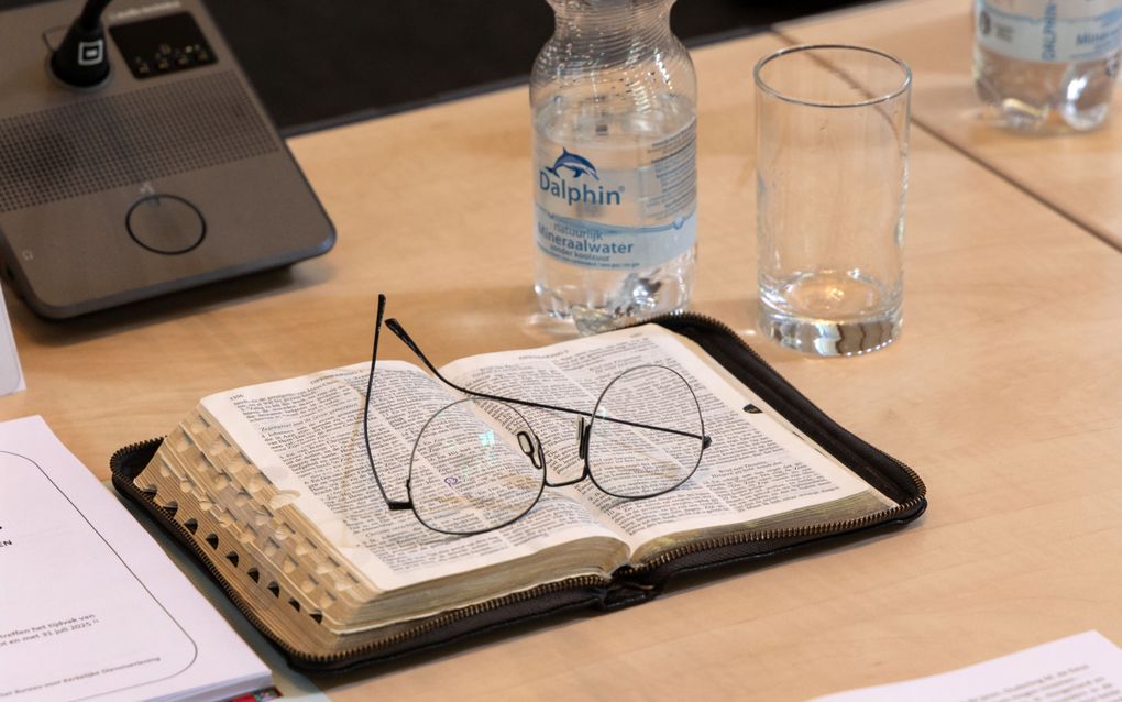 Een opengeslagen Bijbel met een bril erop. Op de tafel verder onder andere een flesje mineraalwater en een glas.