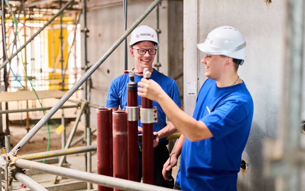 Twee jongemannen met helm zijn bezig met leidingen in een werkplaats.