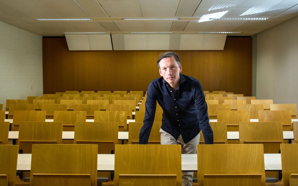 Man van middelbare leeftijd met lichte broek en donkerblauw overhemd staat te midden van een collegezaal met lichthouten stoelen en witte tafels. Hij leunt voorover, met zijn handen steunend op een van de tafels