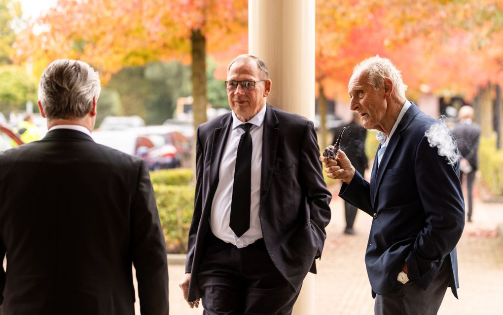 Drie mannen in donkere pakken staan met elkaar te praten, een van hen rookt een pijp.