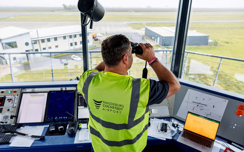 Een man met een geel hesje aan tuurt door een verrekijker vanuit een luchtverkeerstoren over een startbaan van een vliegveld.