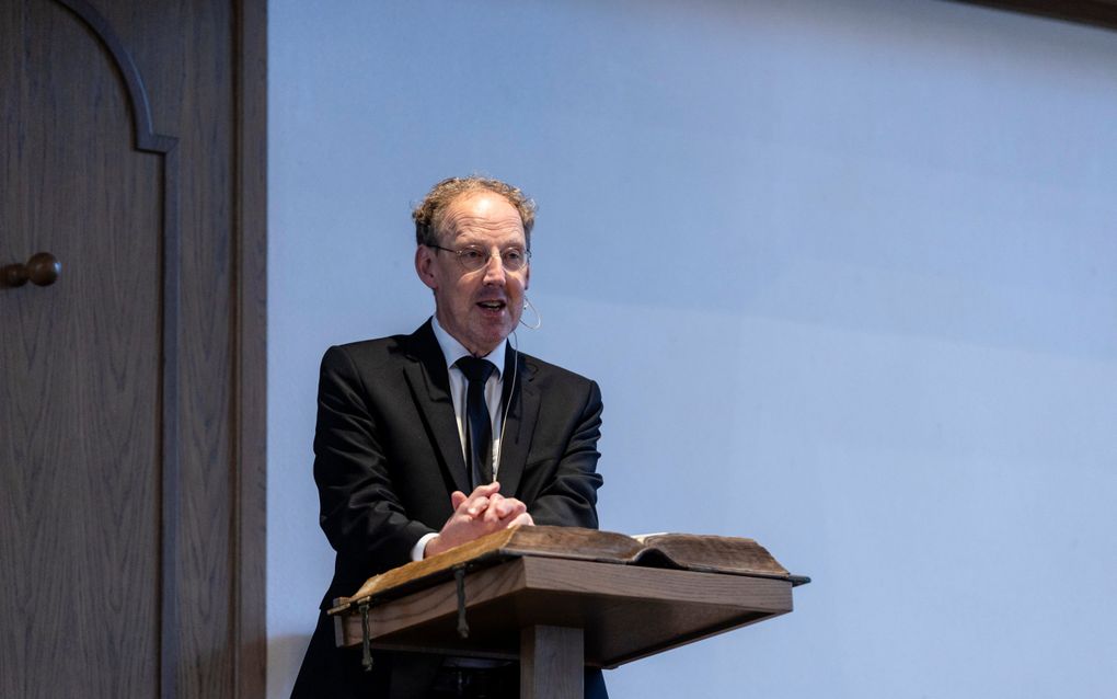 Een man met donkerblond haar, bril en zwart pak spreekt op een kansel.