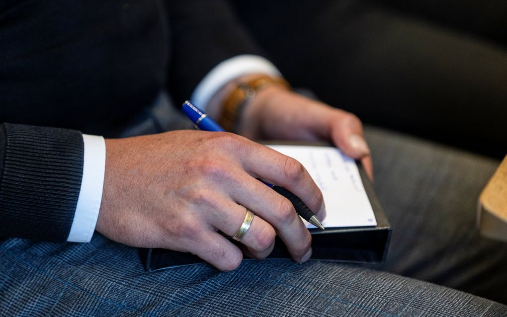 Iemand heeft een notitieblokje op zijn schoot. Ook heeft hij een pen vast.