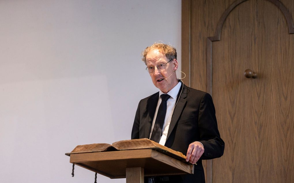 Een man met donkerblond haar, bril en zwart pak spreekt op een kansel.