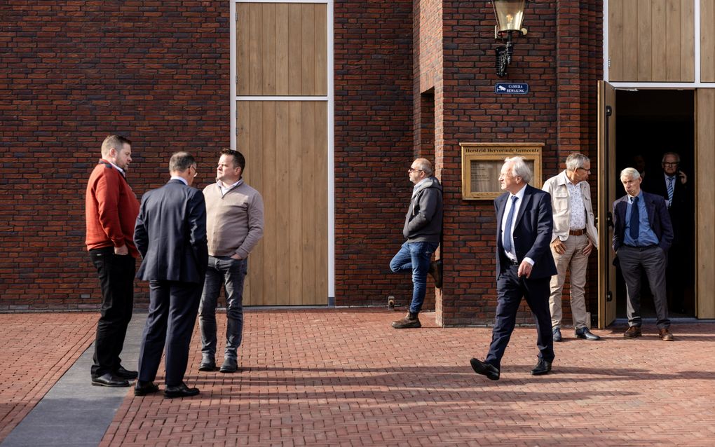 Drie mannen maken een praatje met elkaar op een kerkplein. Enkele mannen lopen de deuropening uit.