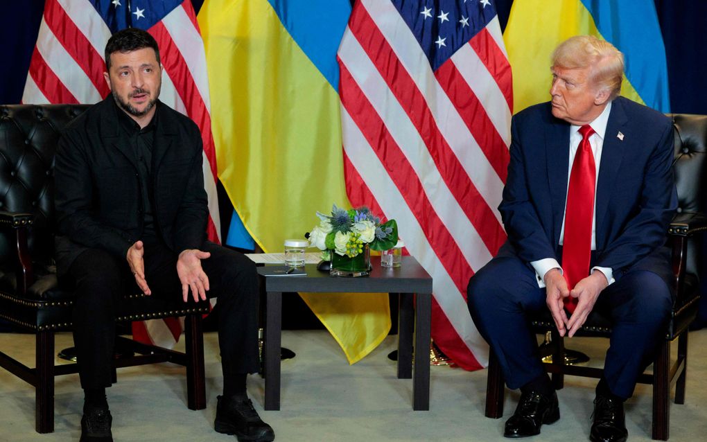 Man in donkere kleding zit naast iemand in formele kleding. Op de achtergrond de Amerikaanse en Oekraïense vlag.