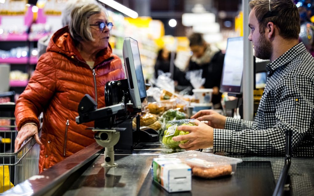 Mevrouw in oranje jas rekent boodschappen, waaronder een bakje druiven, af bij de kassa van een supermarkt.